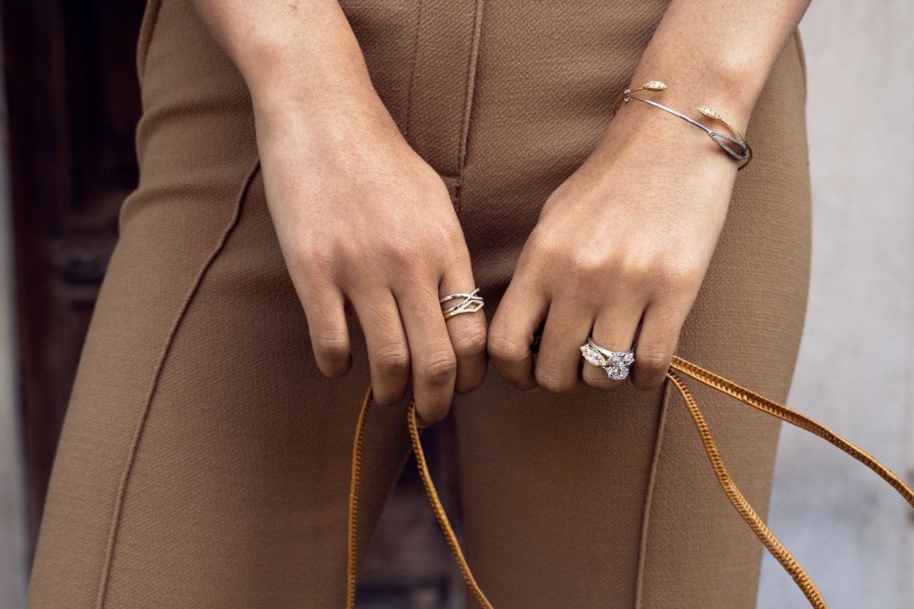 A woman holding the strap of her purse while wearing brown pants, a TACORI fashion ring, bridal stack, and duo of a bangle and cuff in mixed metal tones for each piece by this brand