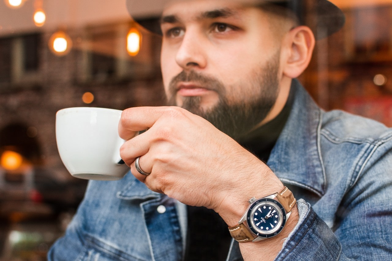 A man in a coffee shop window, holding a mug and wearing a luxurious silver and blue watch with a brown suede strap