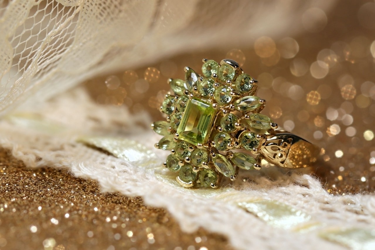 A halo peridot birthstone fashion ring sits under gold glitter and white lace