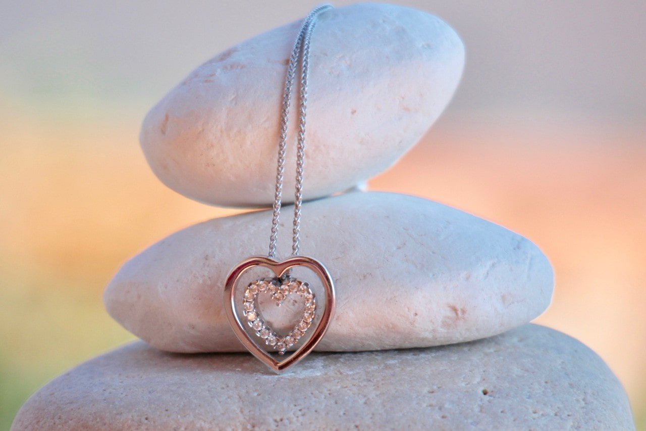 A white-gold heart pendant is draped over a stack of rocks