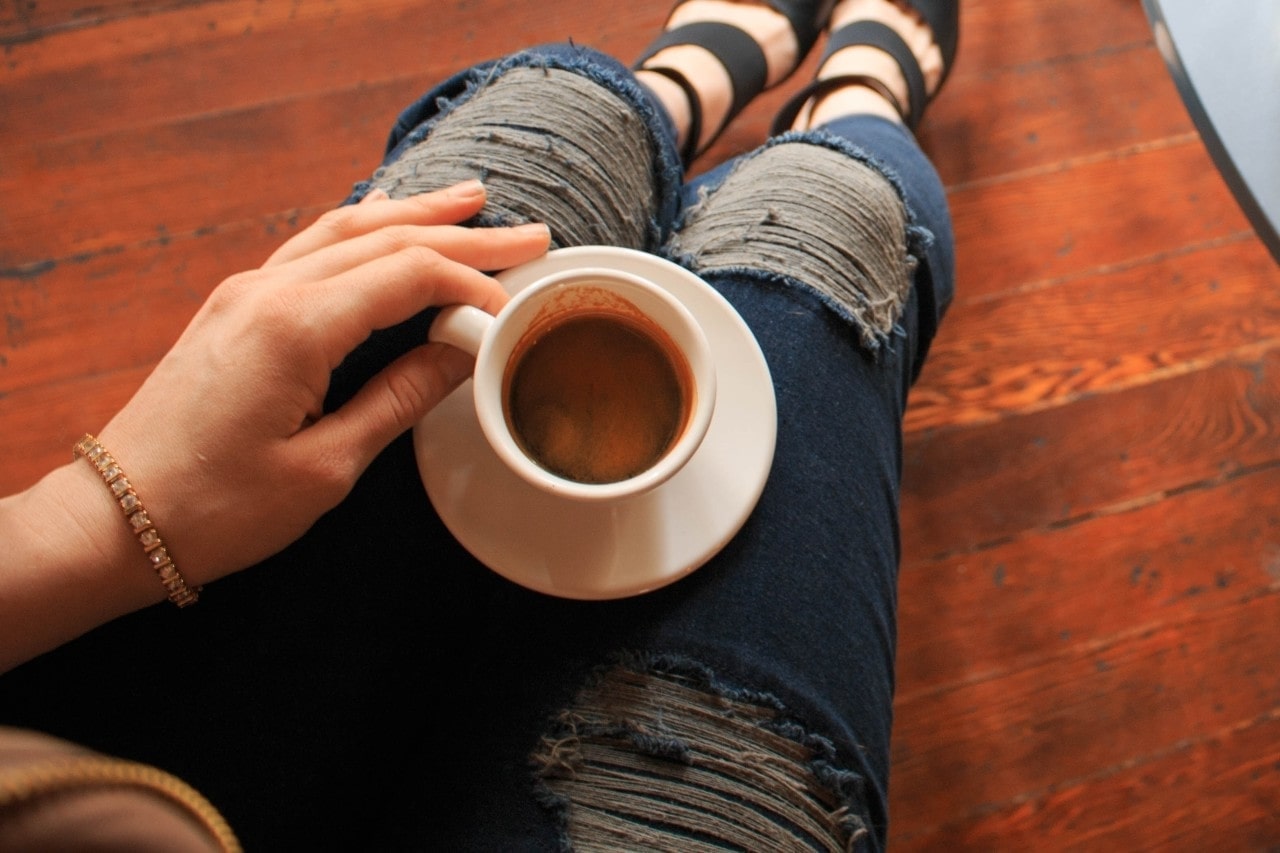 A woman wearing ripped jeans and a gold diamond tennis bracelet sips coffee while sitting on the floor