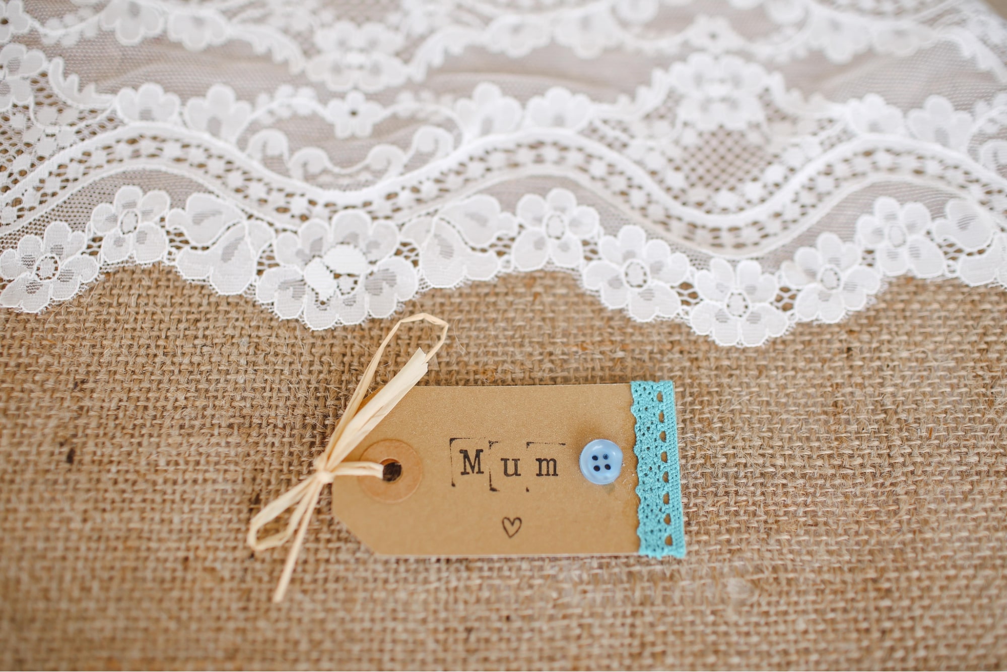 A handmade gift tag for a Mother’s Day gift sits on a burlap runner with white lace