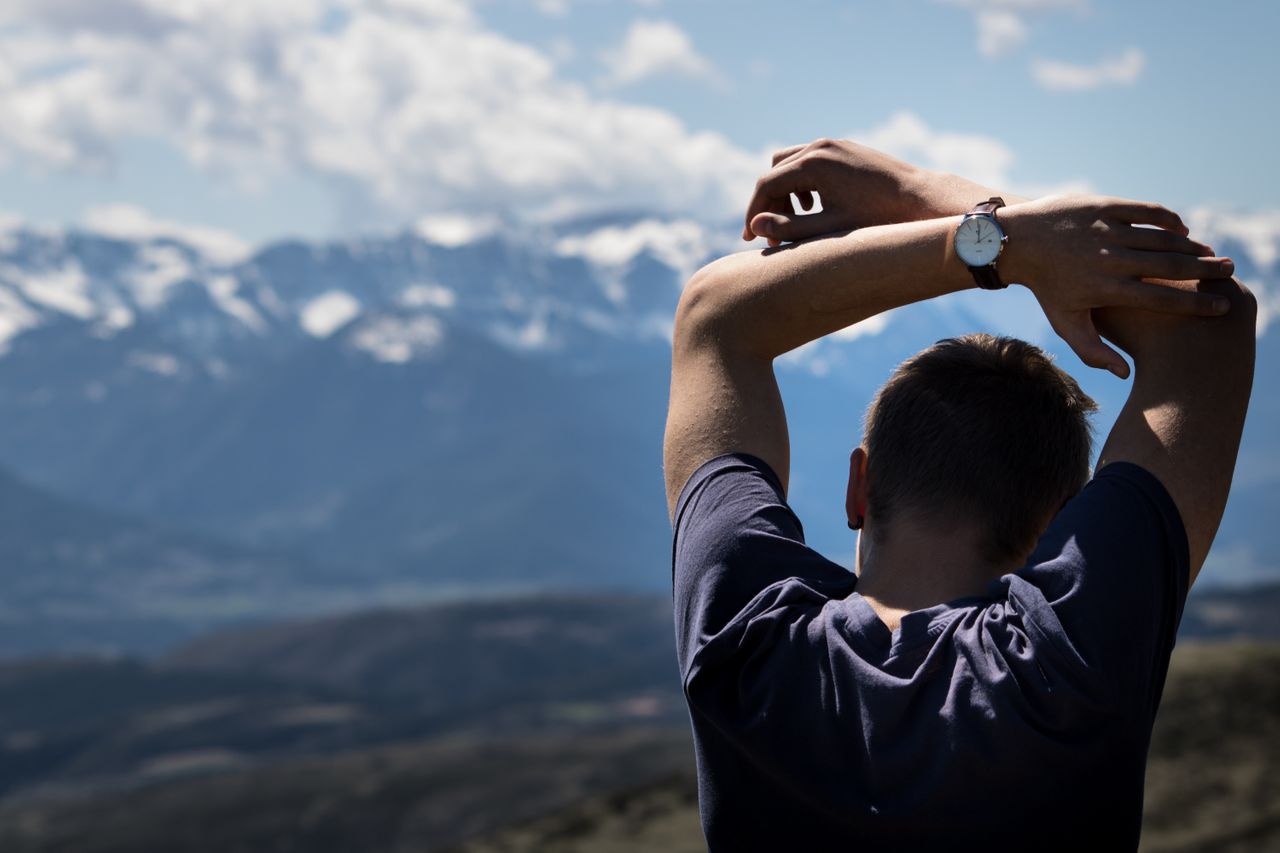 A man wearing a watch raises his hands above his head to catch his breath during a hike