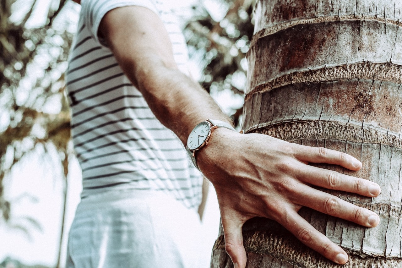 A hiking man wearing a striped shirt places his hand on a tree trunk to show off his watch
