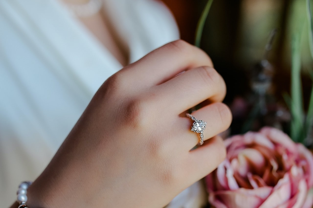 a bride’s hand wearing an engagement ring with a rose in background