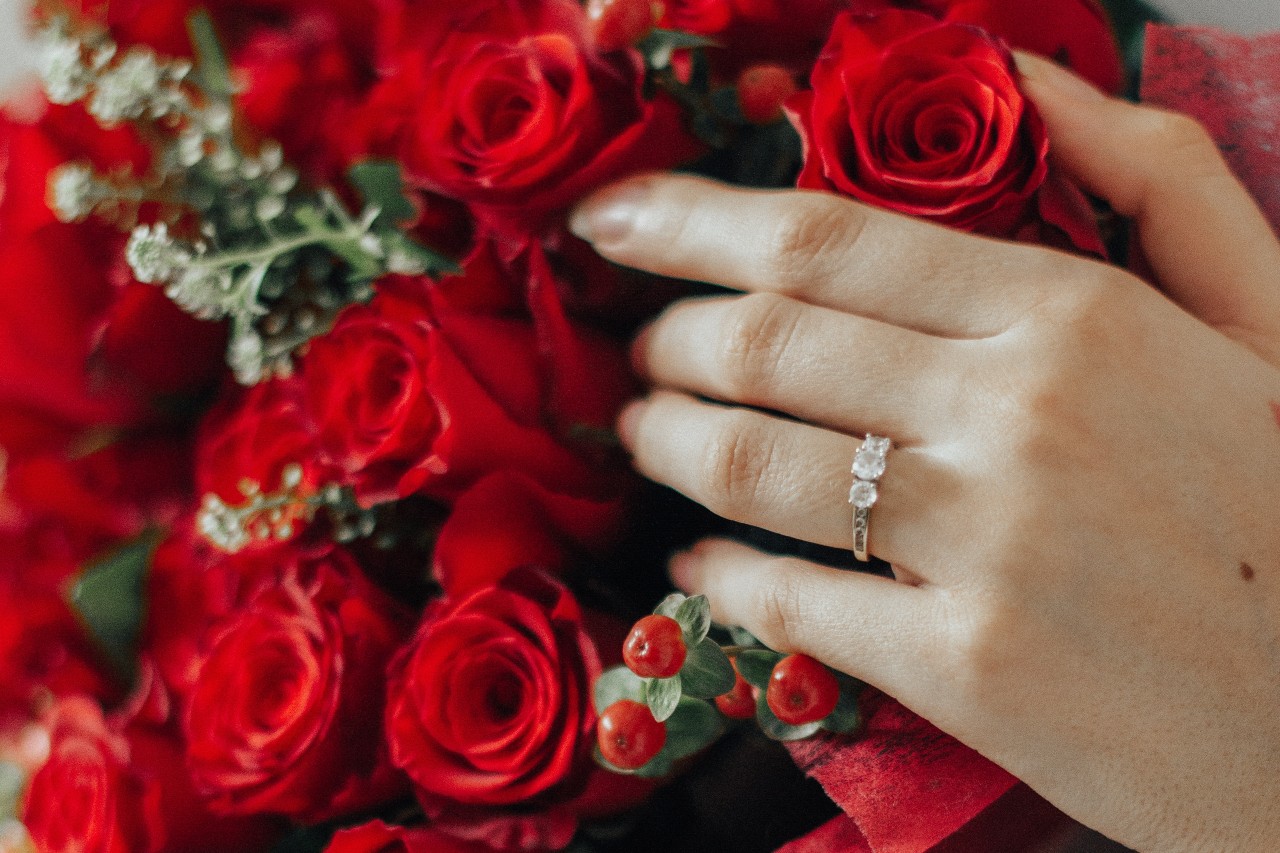 A woman wearing a three-stone engagement ring touches a bouquet of roses.