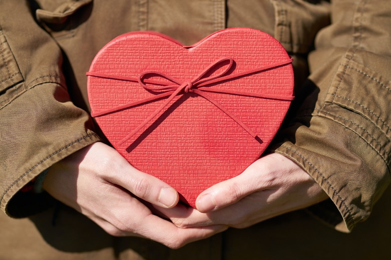 A woman in a brown canvas jacket holds a red heart-shaped box.