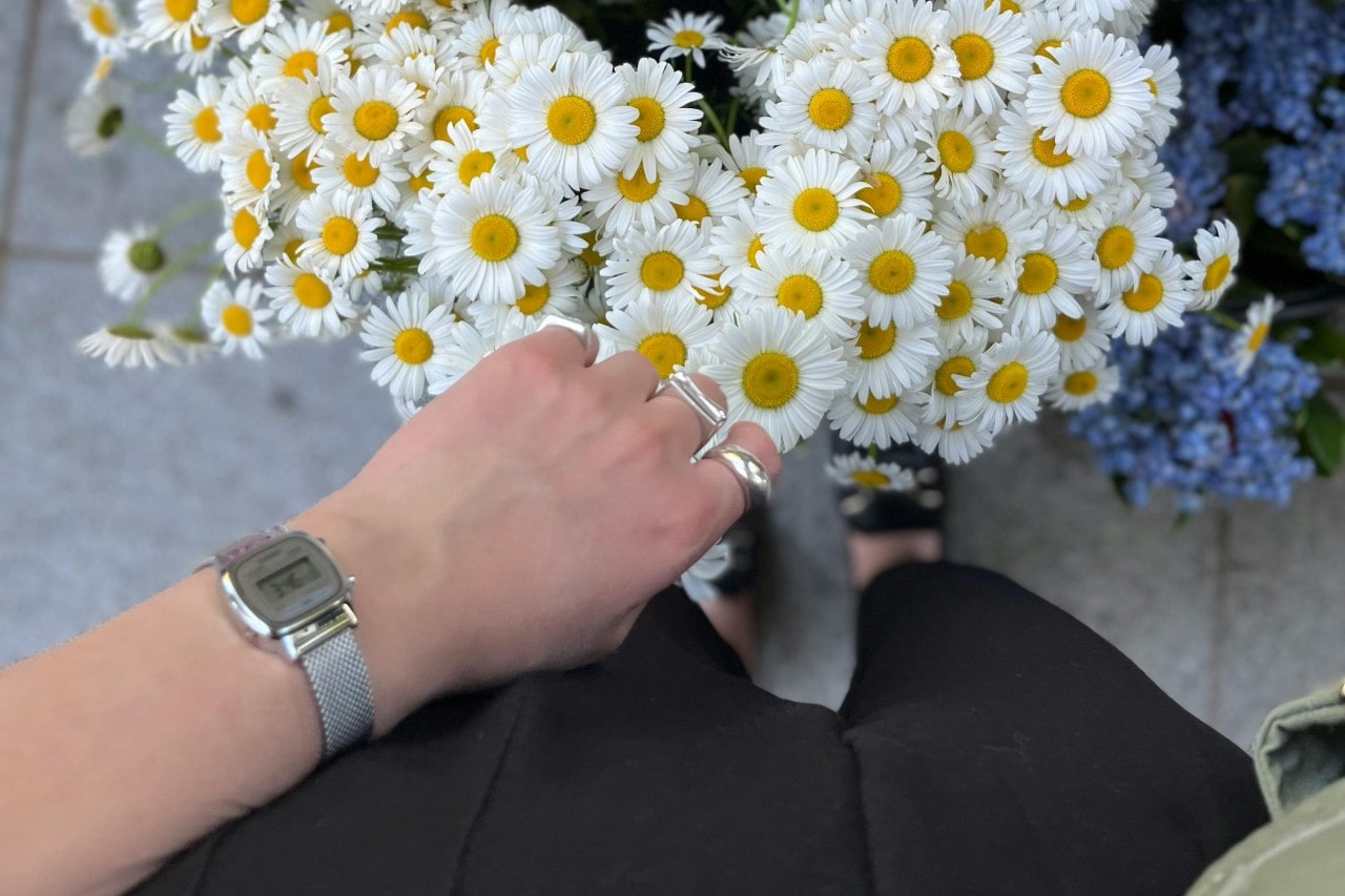 a person standing next to a bunch of flowers, wearing a retro-inspired watch and silver rings