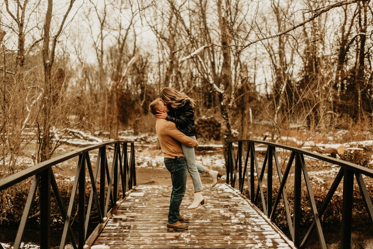 An engaged couple kissing on a bridge in a park dusted with snow.