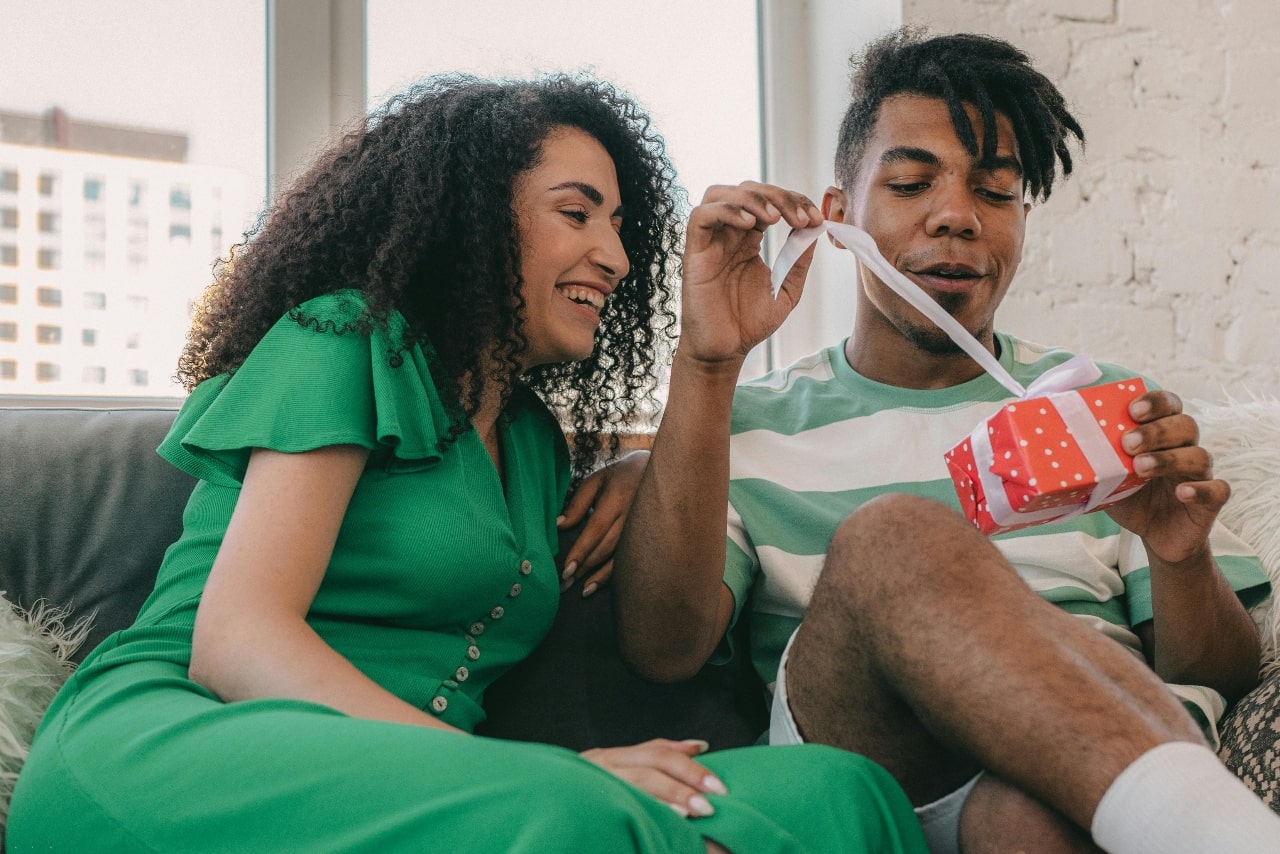 A woman in the green with curly hair sits on a couch and watches how a man in a striped shirt opens a gift box.