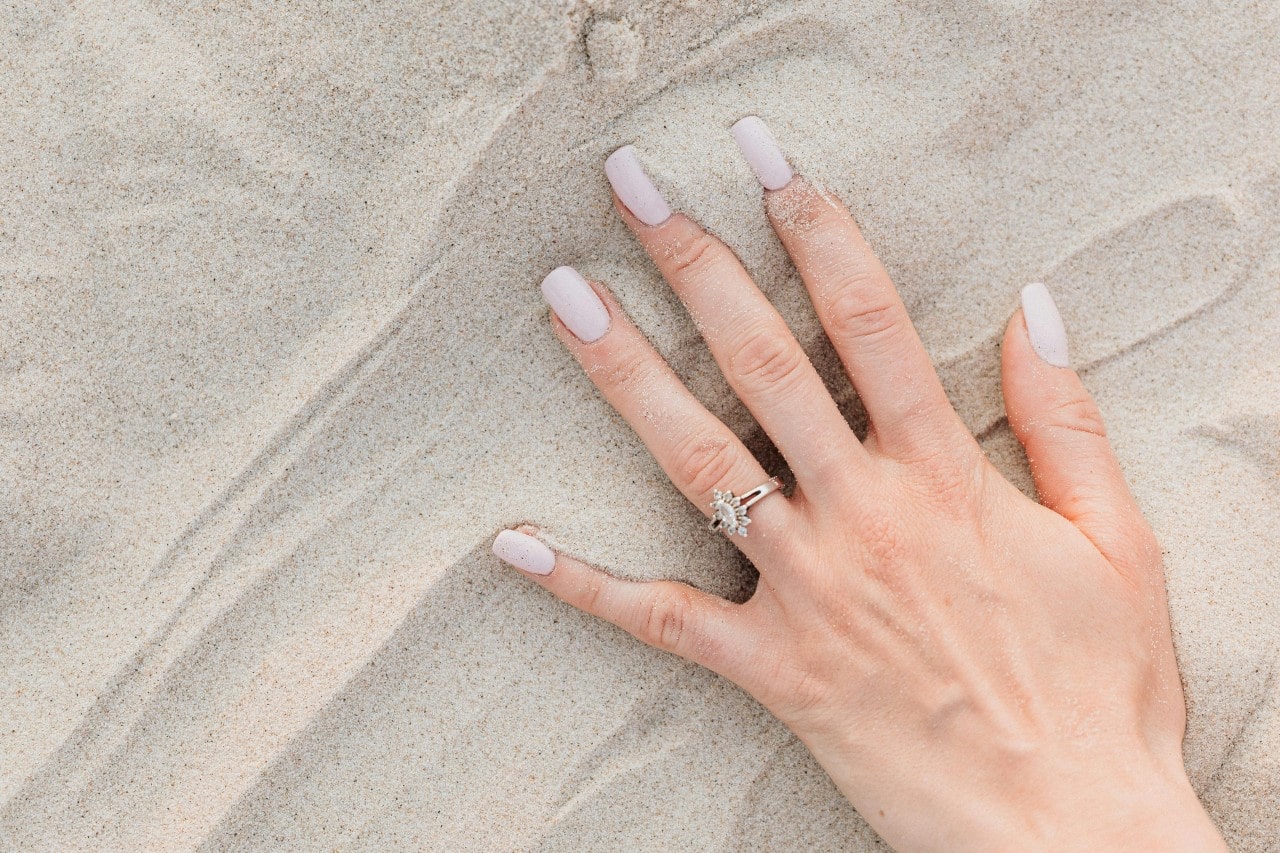 a woman’s hand in the sand, wearing a marquise diamond halo engagement ring