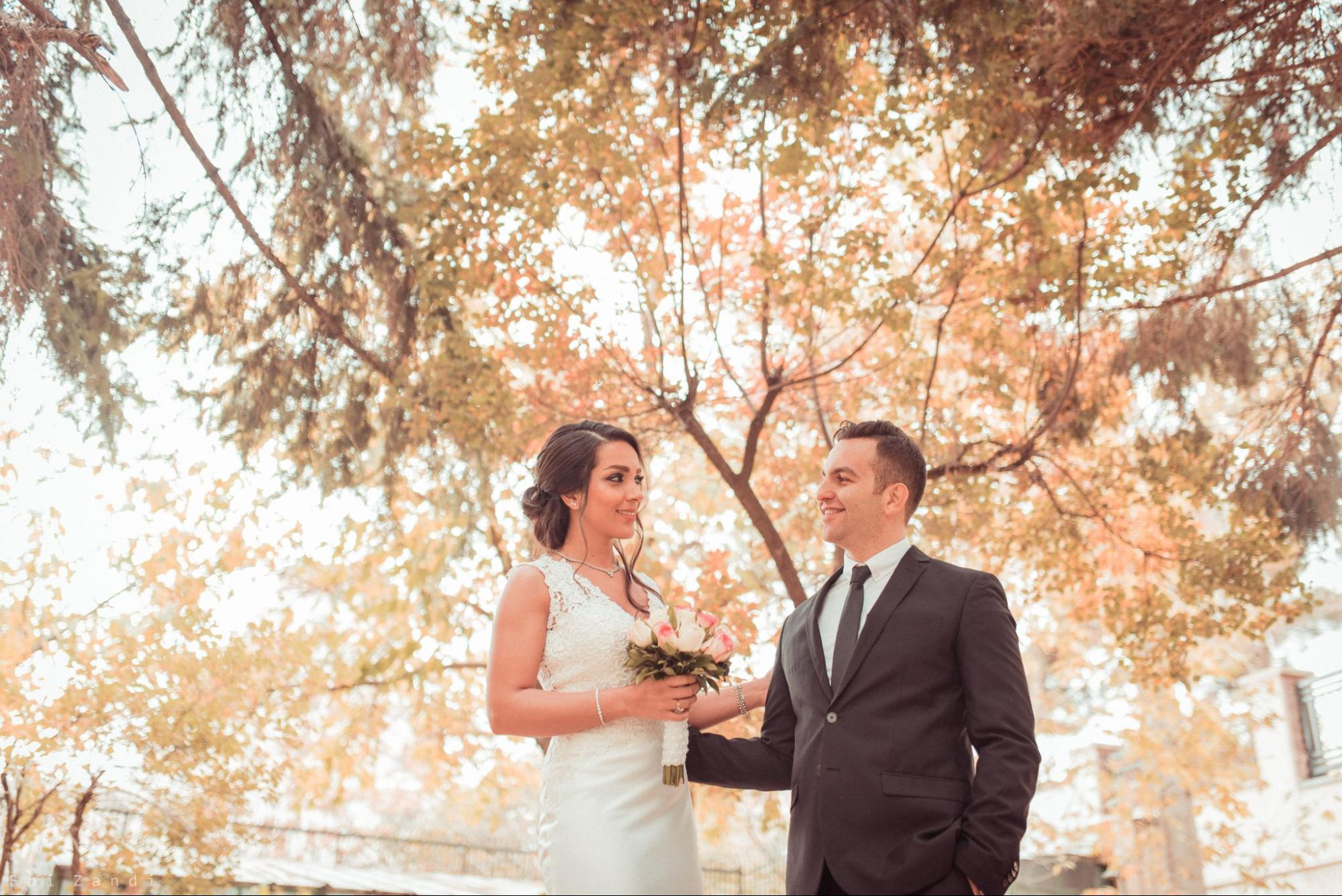 A newly married couple smiling at each other under trees in the fall.