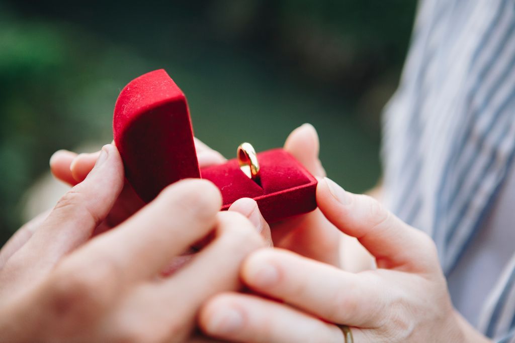 A person presents their partner with a simple gold wedding band in a red box.