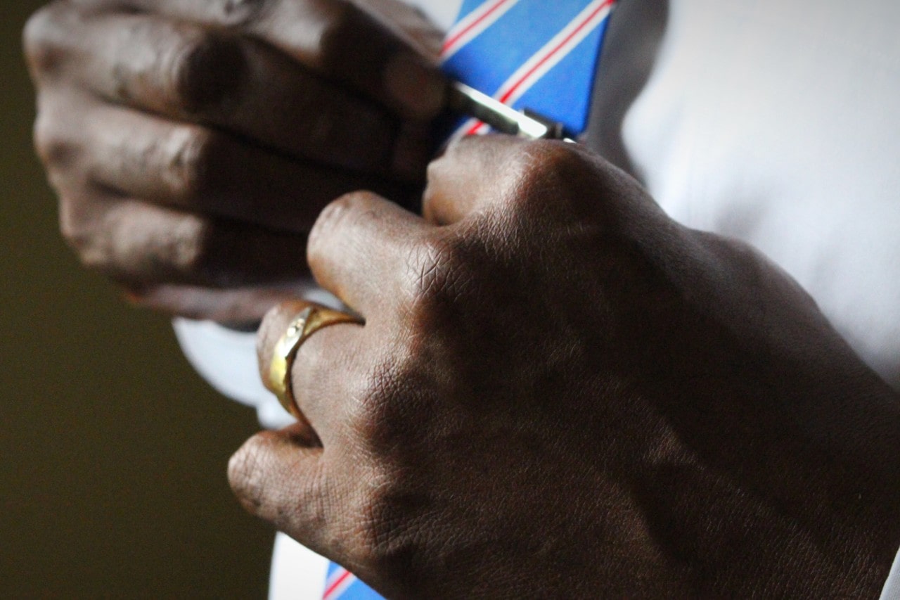 A close-up of a well-dressed man tying a tie, and wearing a gold wedding band.