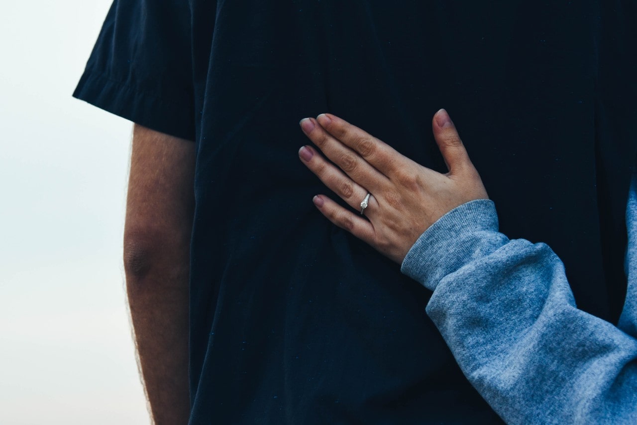 woman’s hand resting on her partner’s back, wearing a white gold engagement ring