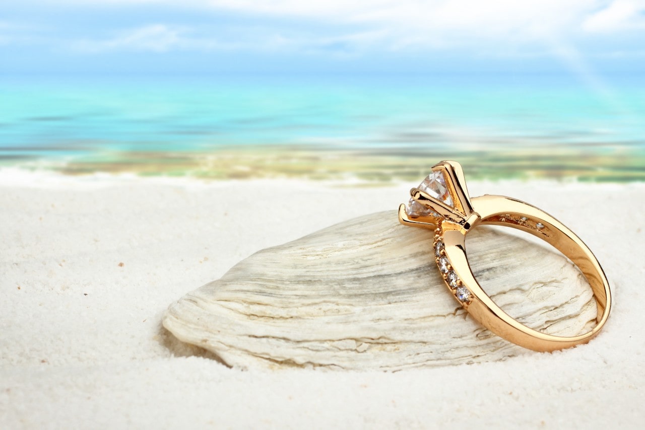 a yellow gold side stone engagement ring resting on a shell on a beach