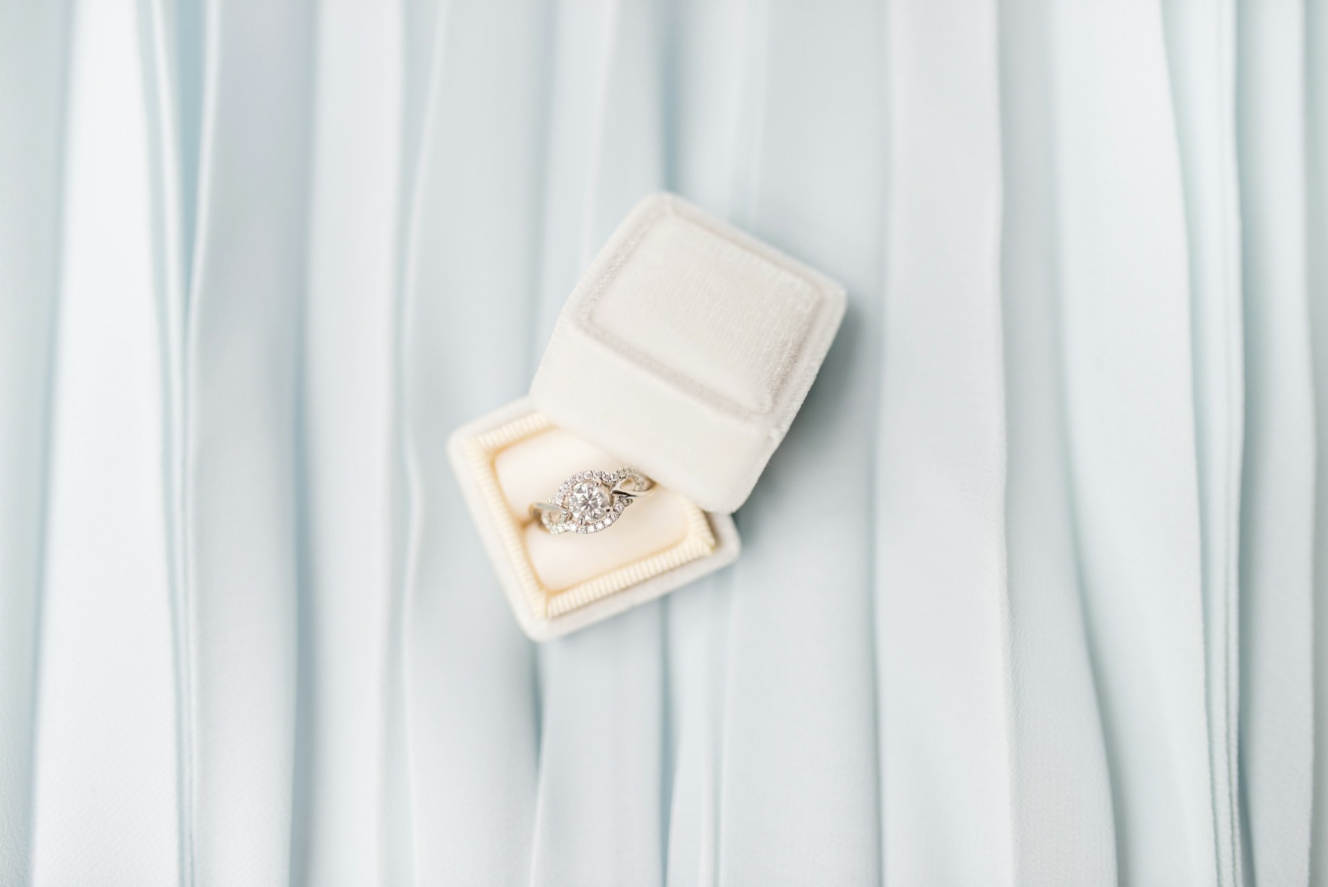 A unique halo engagement ring in a white box on a light blue fabric background.