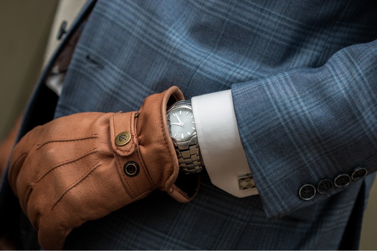 A close-up of a silver watch with a clean white dial, worn under a blue checkered suit jacket and accessorized with a brown leather glove