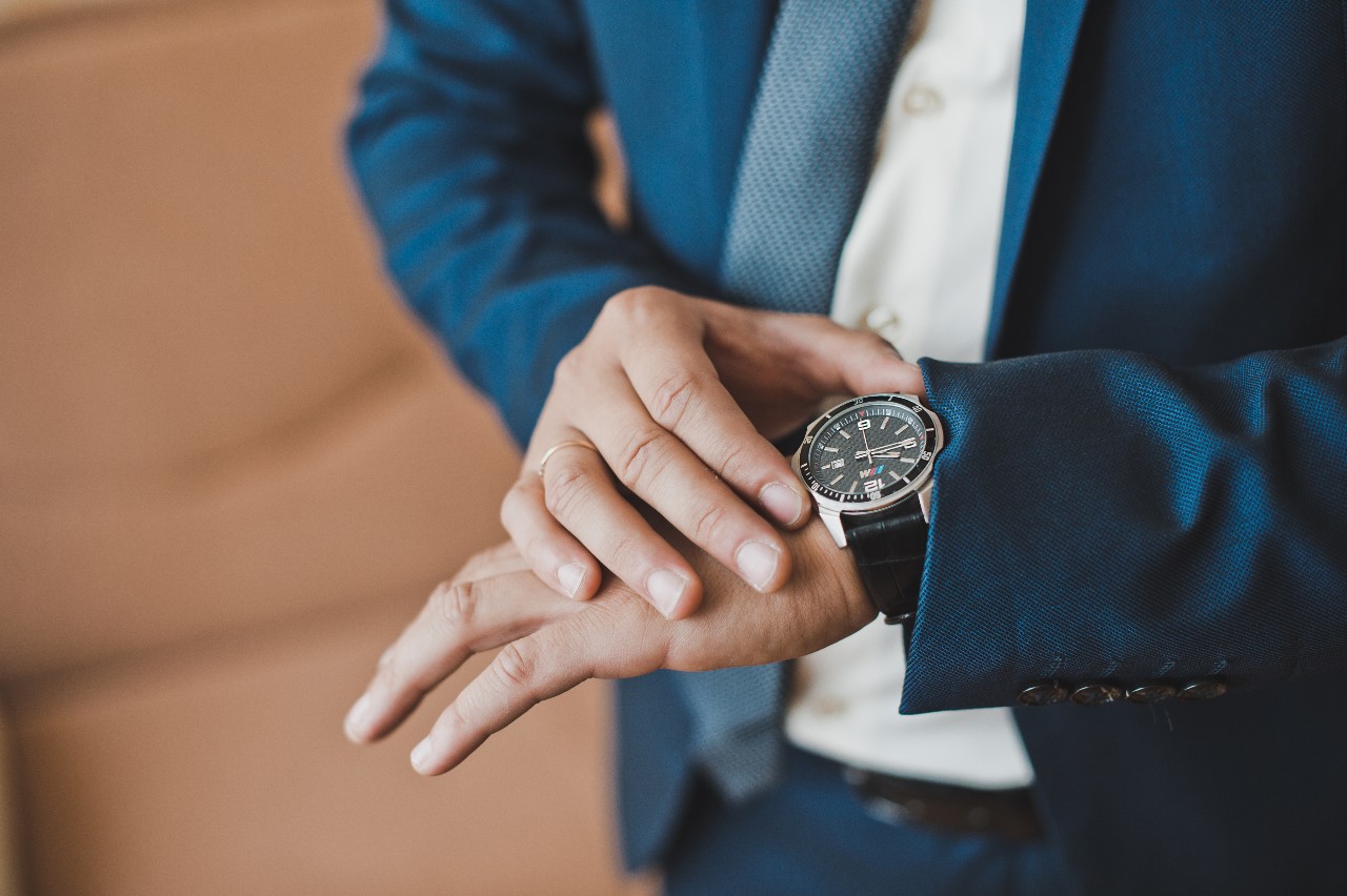 A dark-faced wristwatch with bold white hour markers and a black leather strap, worn by a man in a navy suit with a white shirt and blue tie