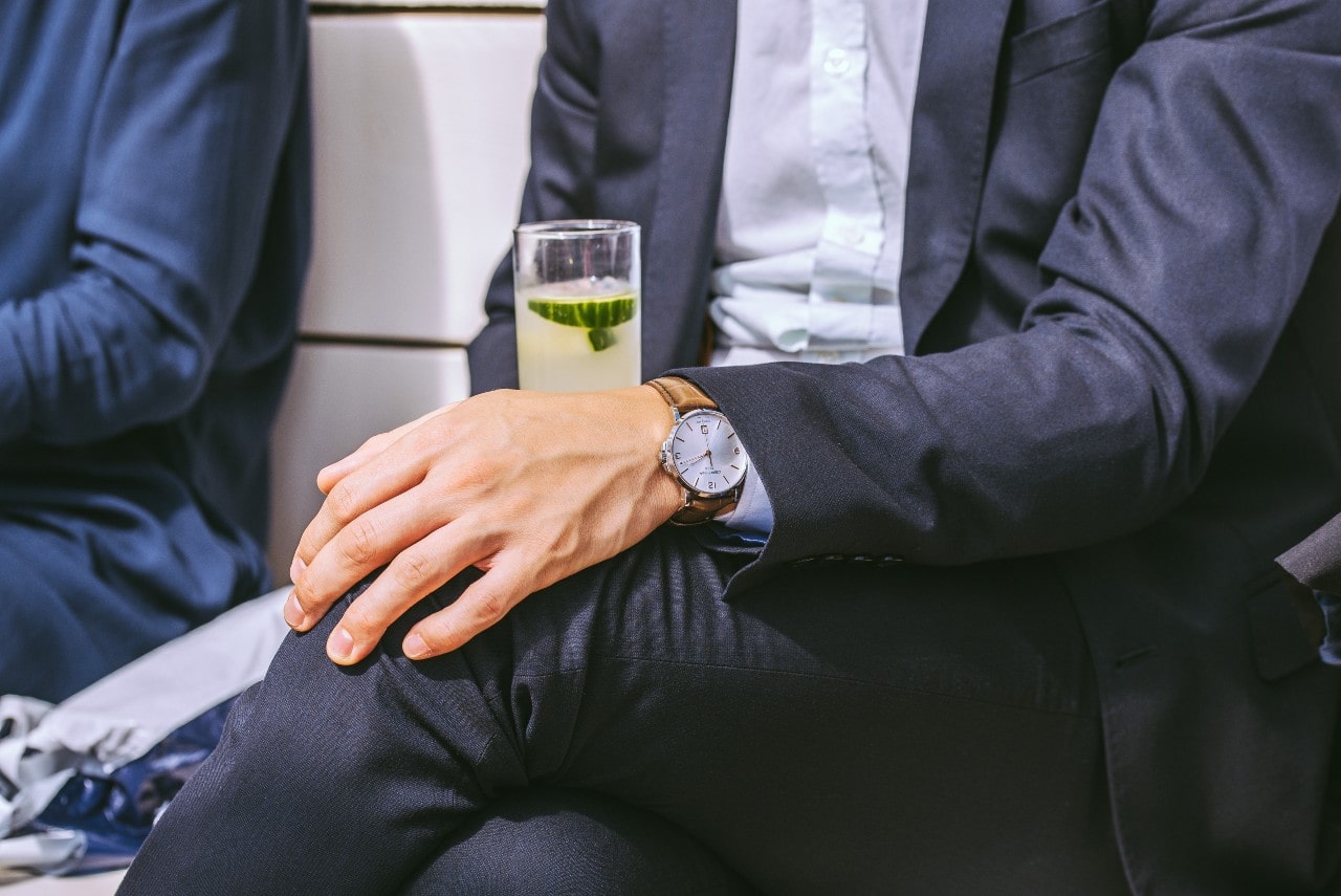 A man in a navy suit with a stylish watch on his wrist rests his hand on his leg while holding a glass of lemonade with cucumber slices